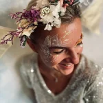 Auf dem Bild sieht man eine Frau mit einem Blumenkranz im Haar. Sie wird von einem Blüten Make Up geschmückt. Sie trägt ein funkelndes Pailetten Oberteil. Glitzerbar Junggesellenabschied