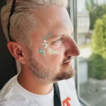 Hier sieht man einen Mann von der Seite. Er hat ein Glitzer Make Up am rechten Auge und schaut aus dem Fenster. Facepainting für Festivals