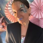 Auf diesem Bild sieht man eine hübsche Frau mit einem Festival Make Up auf der linken Gesichtshälfte. Sie schaut lächelnd direkt in die Kamera und trägt blauen Lippenstift. Sie steht vor einem pastellfarbenen Hintergrund. Event Schminken Glitzerbar Rhein-Main-Gebiet