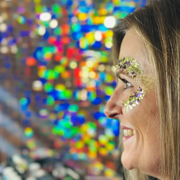 Lächelnde Frau schaut in den Spiegel. Man sieht sie von der Seitenansicht lächeln. Sie trägt ein Glitzer Augen Design in den Farben gold und lila. Glitzerbar für Firmenfeiern