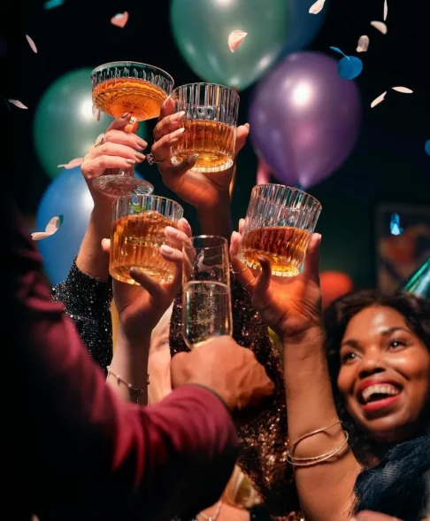 Hier sieht man feiernde Menschen, die sich zu prosten. Im Hintergrund sind bunte metallic Luftballons und eine Frau lächelt. Teambuilding Frankfurt Corporate Events