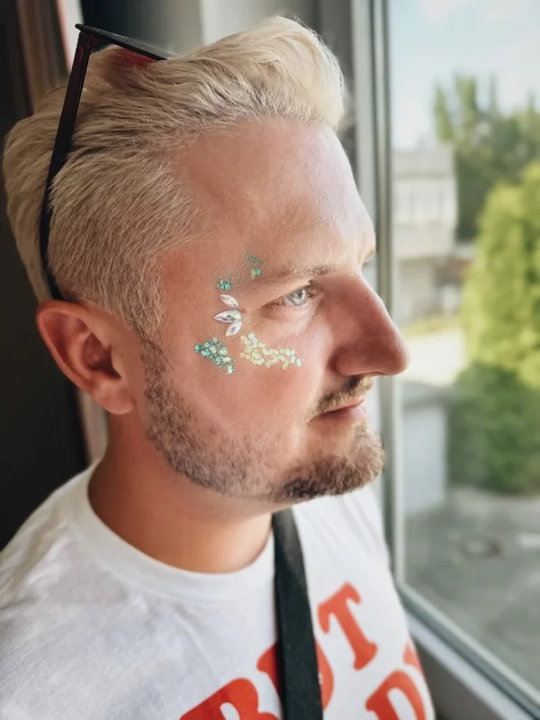 Mann mit blondem Haar und gepflegtem Bart trägt glitzernde Gesichtskunst mit schimmernden Steinen und Glitzerpartikeln auf der Wange