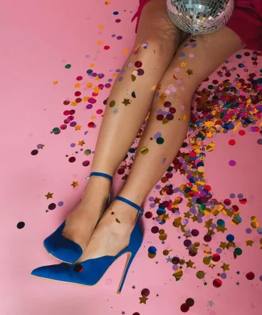 Elegante Beine in blauen High Heels, umgeben von buntem Konfetti und einer glitzernden Discokugel auf rosa Hintergrund.